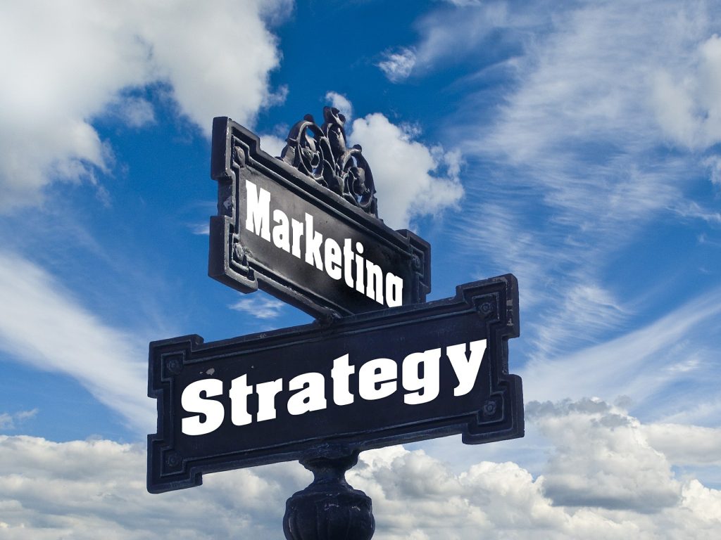 Blue old-fashioned street sign on a blue sky background with the word "marketing" pointing one way and the word "strategy" pointing the other way.
